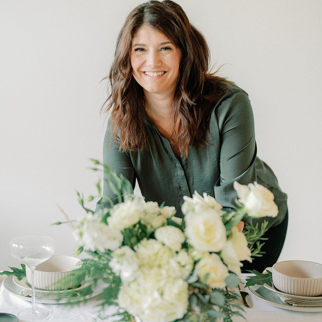 A smiling woman with shoulder-length brown hair sits behind a white table setting decorated with a large bouquet of white flowers, including roses and hydrangeas. She is wearing a dark olive green blouse against a plain white background.