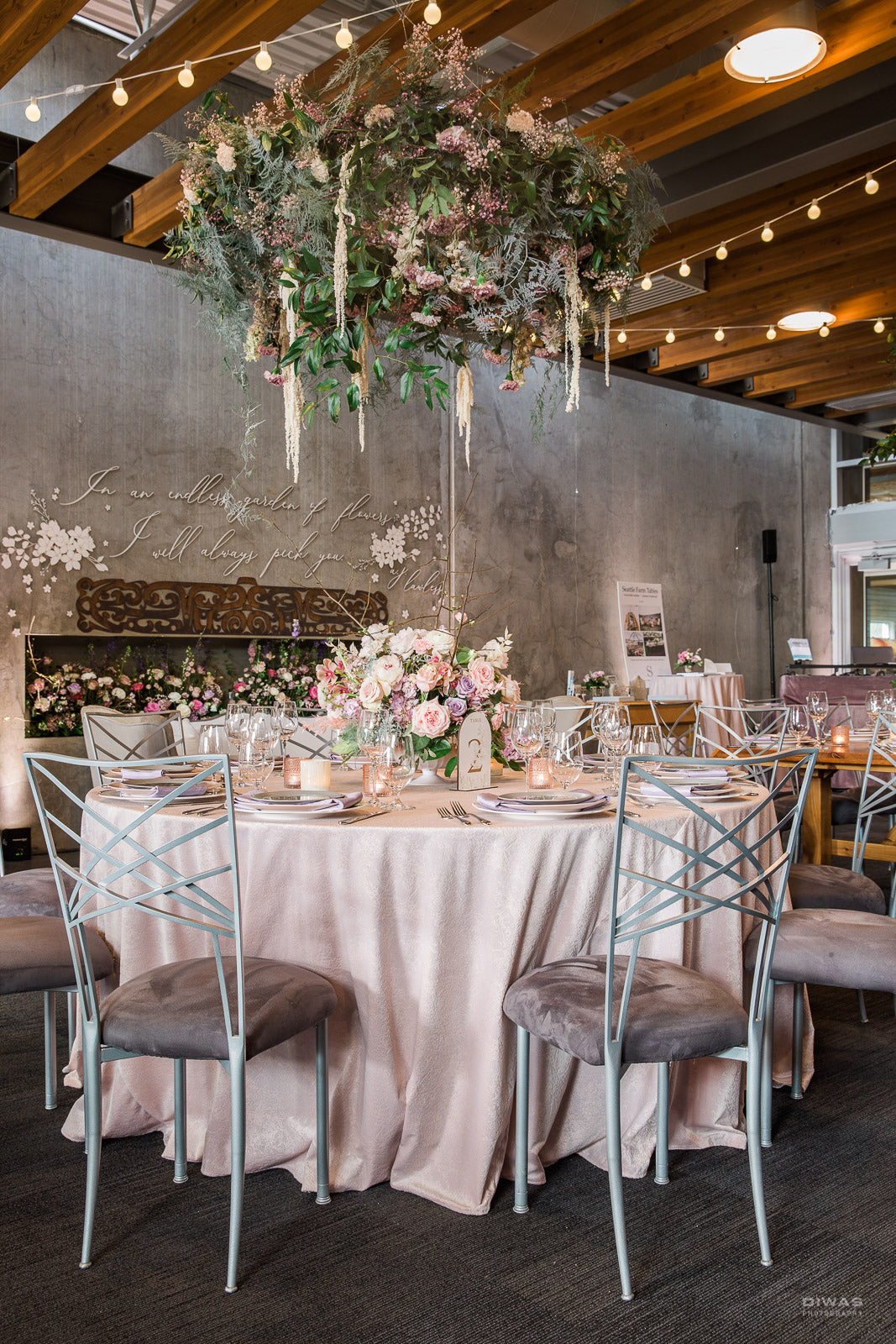 A styled event reception table setup in a rustic indoor venue with exposed wood ceiling beams and string lights. The main subject is a round table covered in a blush pink tablecloth, surrounded by silver X-back chairs with gray upholstered seats.