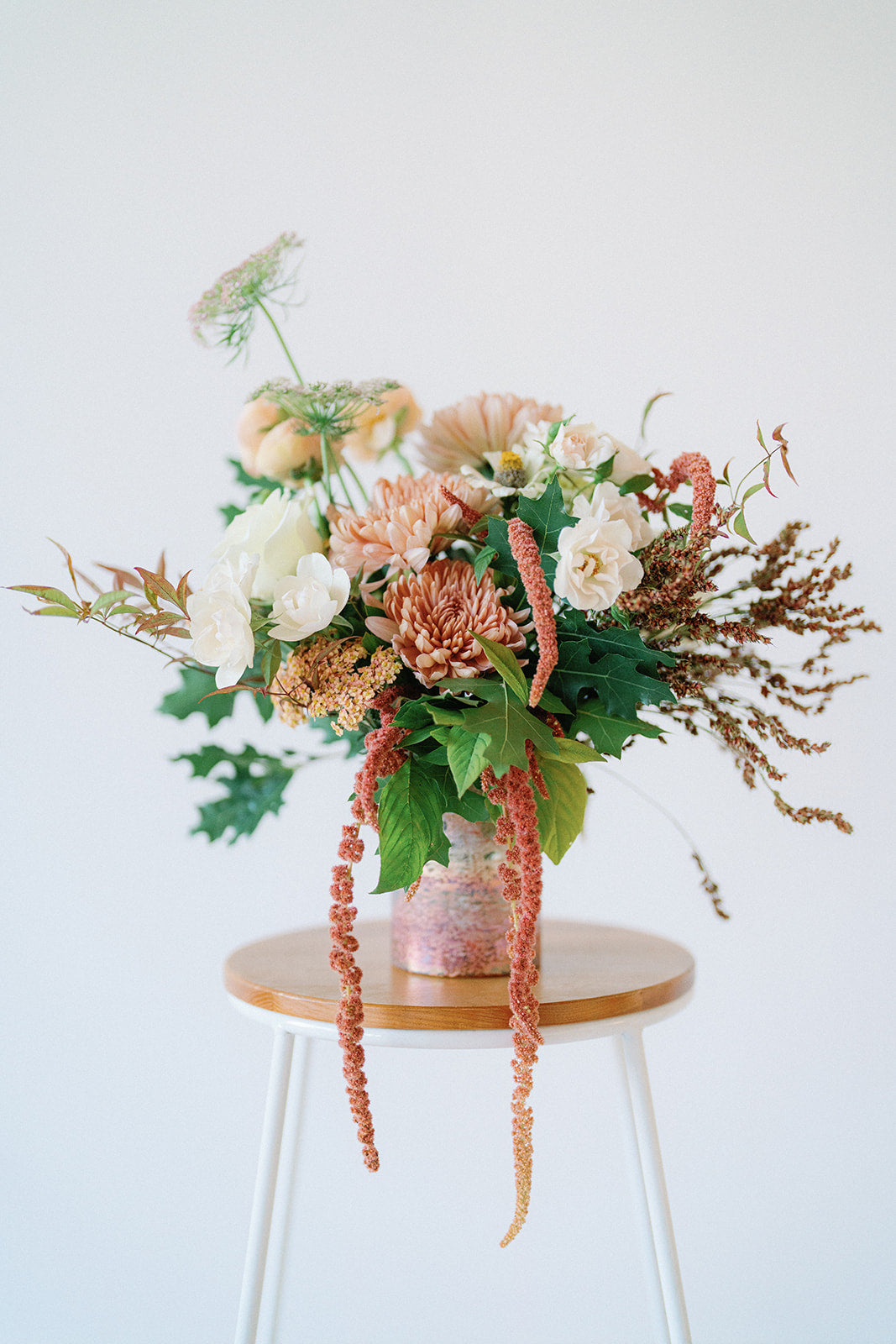 A rustic floral arrangement in a pink textured vase, displayed on a wooden-topped stool against a clean white background.